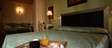 Habitación de hotel con cama, mesita de noche, lámpara, cuadro enmarcado en la pared y bandeja de desayuno con cruasanes y jugo de naranja.