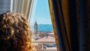 Vista a través de una ventana con cortinas, mostrando un edificio con una torre de reloj y montañas al fondo.