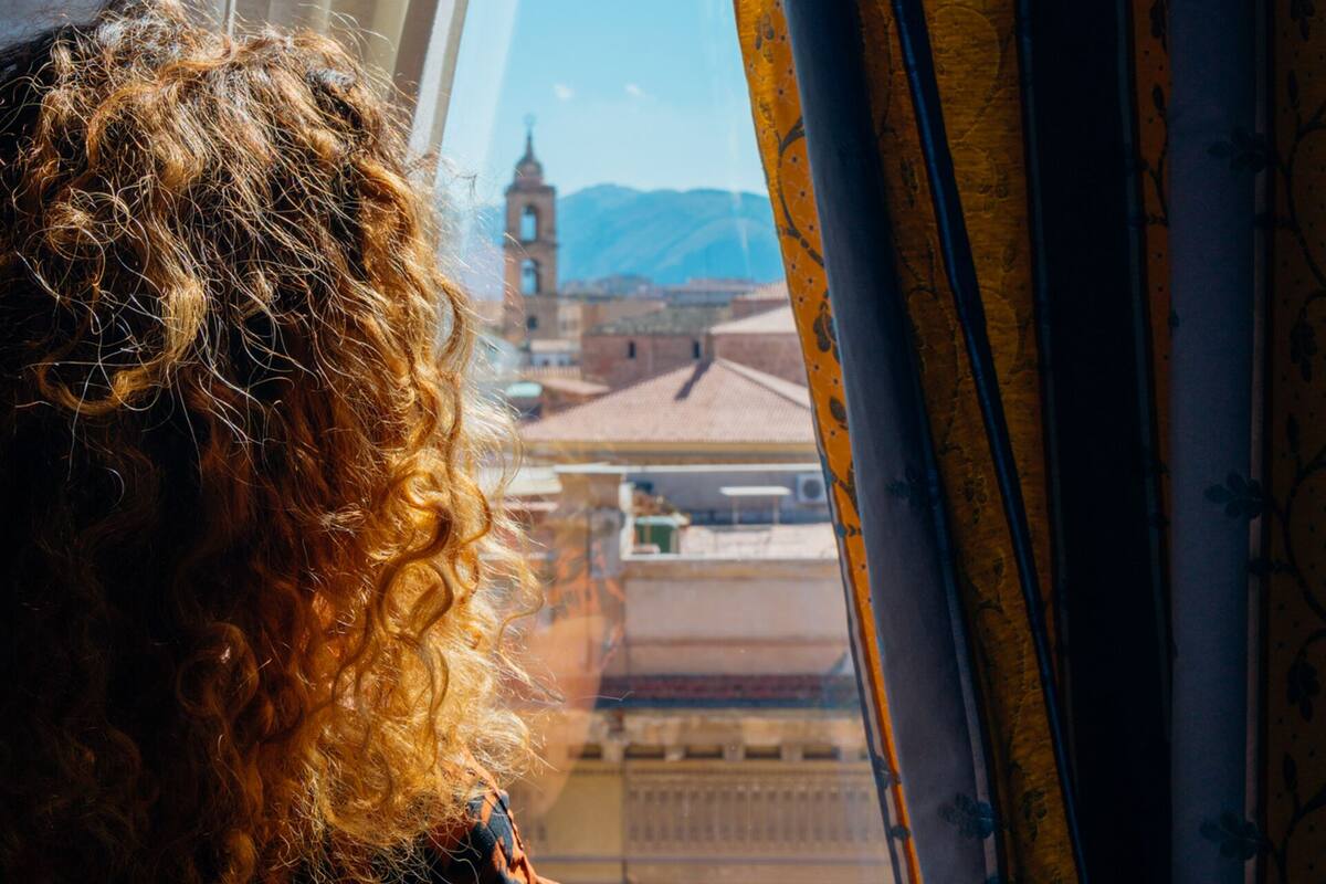 A view through a window with curtains, featuring a building with a clock tower and mountains in the background.