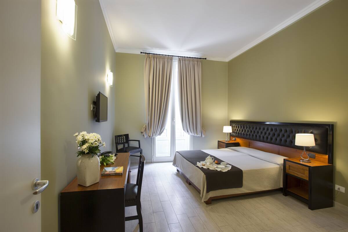 A hotel room with a bed, bedside tables, a desk with a chair, a vase of flowers, and a television on the wall.