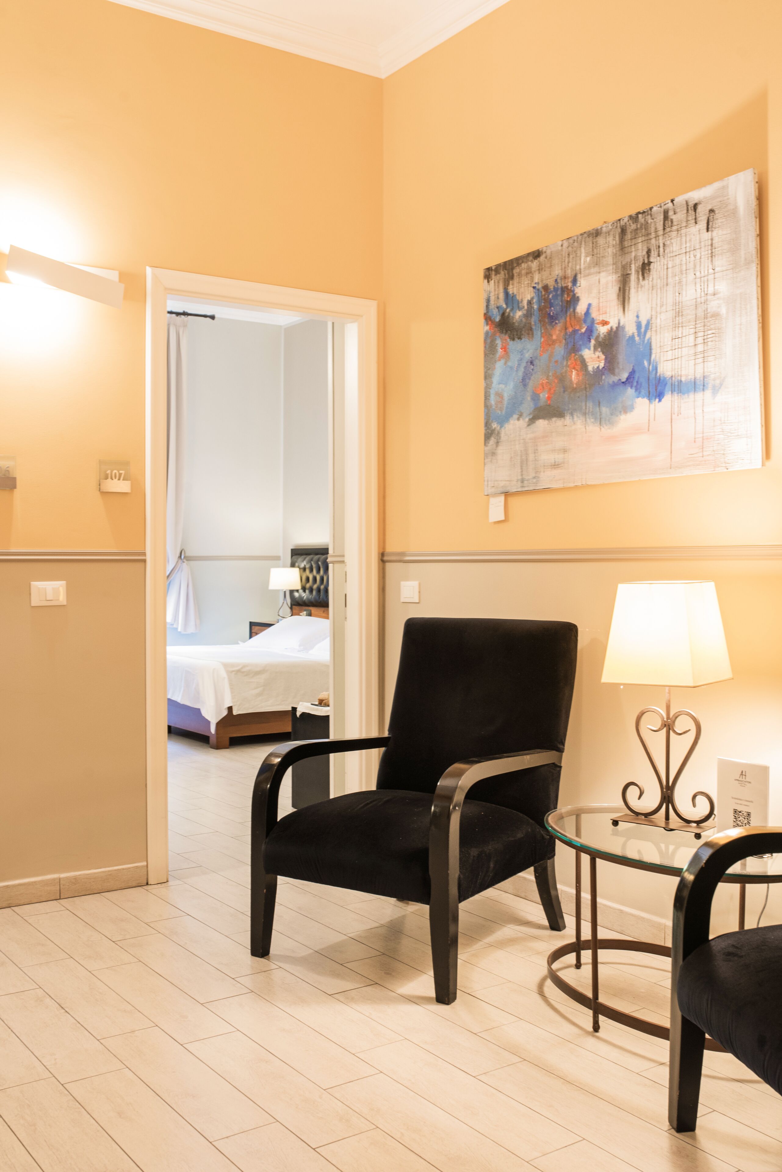 A hotel room with a bed, a black chair, a glass table, and a painting on the wall.
