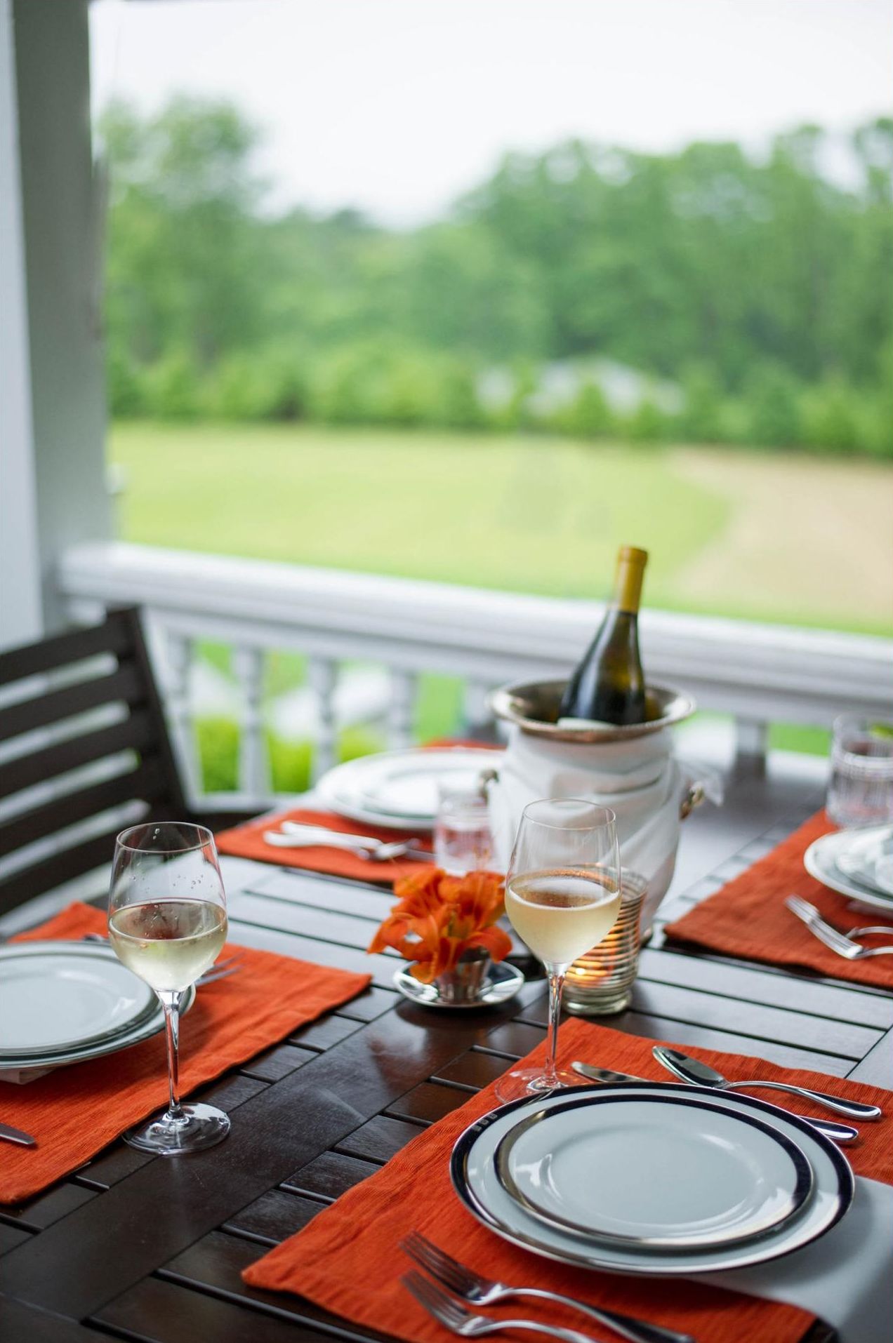 A dining table set with plates, glasses, and a bottle of champagne, with a view of a green landscape.