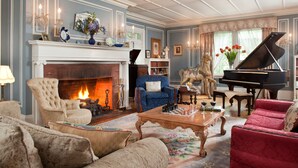 A traditional living room with a fireplace, a grand piano, plush seating, and a bookshelf. - Hampton Terrace Inn (Lenox)