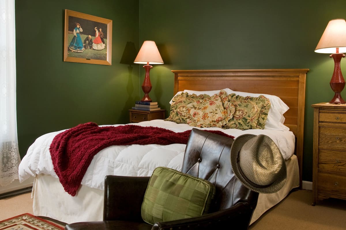 Un letto rifatto con una coperta rossa, un cuscino verde e un cappello di paglia su una sedia. Ci sono due comodini in legno con lampade e un quadro appeso al muro.