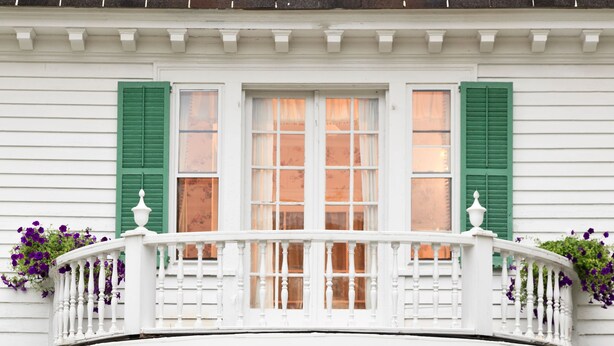 A white house with a balcony, green shutters, and a front door with a decorative glass panel. There are two large flower pots on the steps. - Hampton Terrace Inn (Lenox)