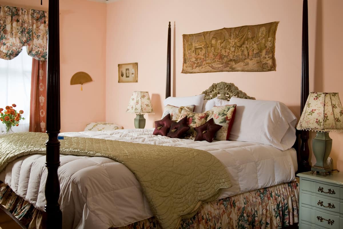 Una camera da letto con un letto a baldacchino, tende a motivi floreali, una lampada in stile vintage e un quadro appeso al muro.