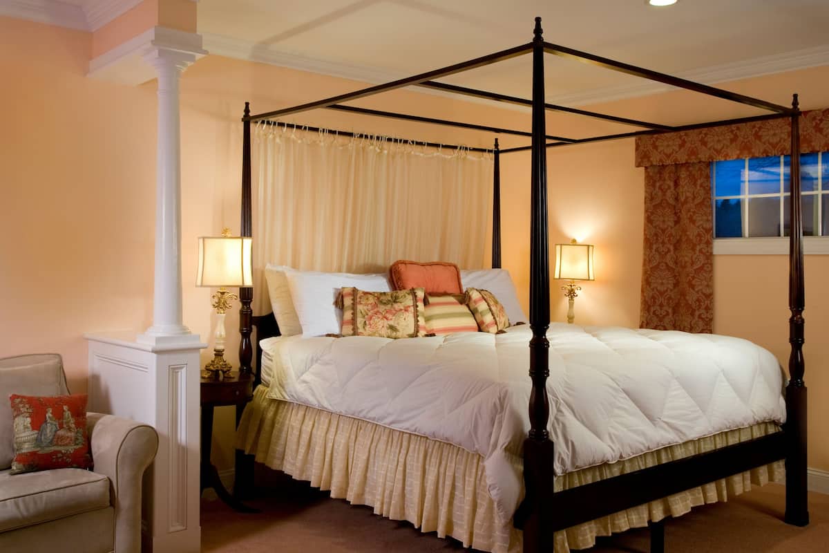 Un letto a baldacchino con tende, un comodino con una lampada e una sedia con un cuscino.