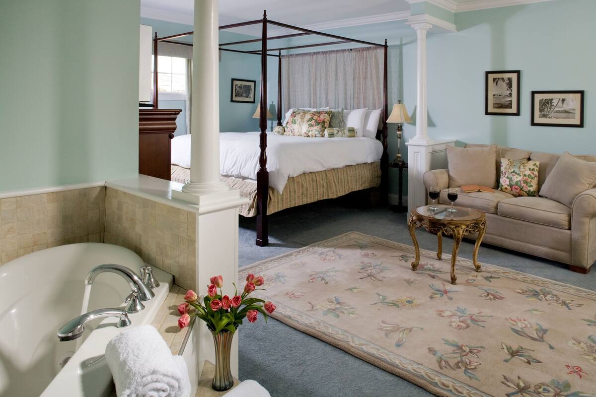 Una camera da letto con un letto a baldacchino, una zona salotto con divano e un tavolino, un'ampia finestra e un bagno con vasca e lavabo.