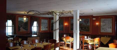 Ein Restaurant mit Holztischen und -stühlen, gerahmten Kunstwerken an den Wänden und einer Kristallleuchter.