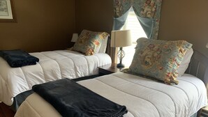 A hotel room with two beds, each with a white duvet and floral-patterned pillows. There is a lamp on a bedside table and a framed picture on the wall.