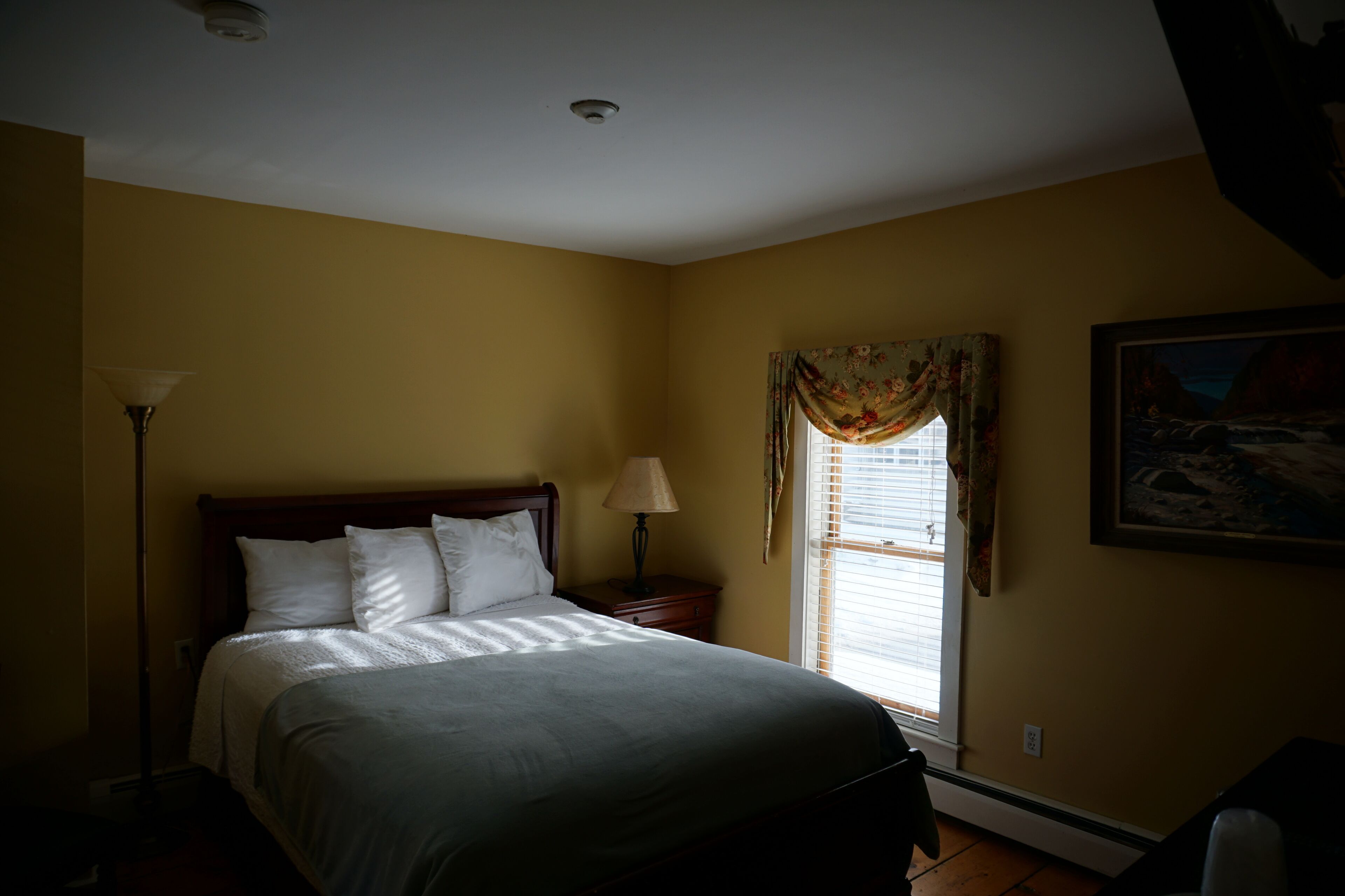 A bedroom with a bed, a nightstand, a lamp, a window with curtains, and a painting on the wall.