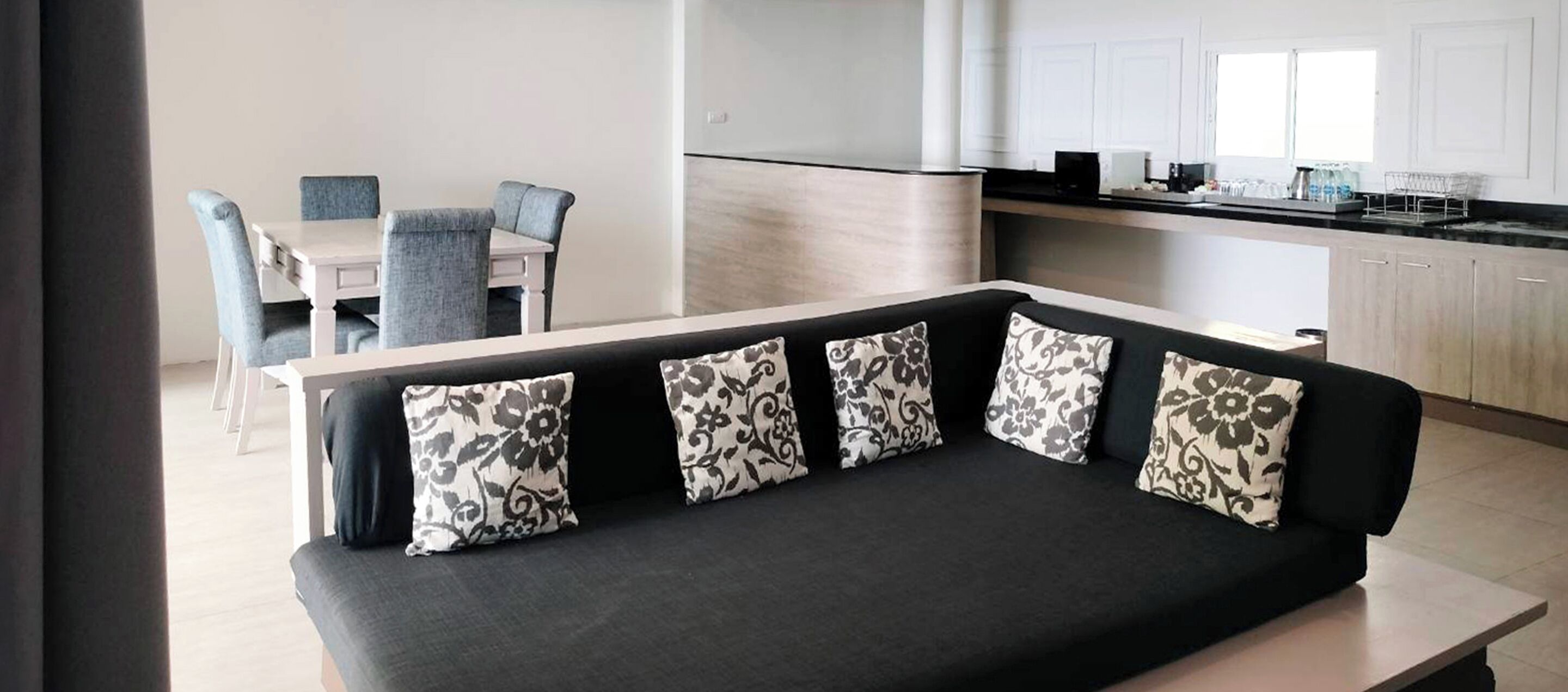 Un espace de vie moderne avec un canapé noir, des coussins à motifs floraux, une table à manger avec des chaises, et une cuisine en arrière-plan.