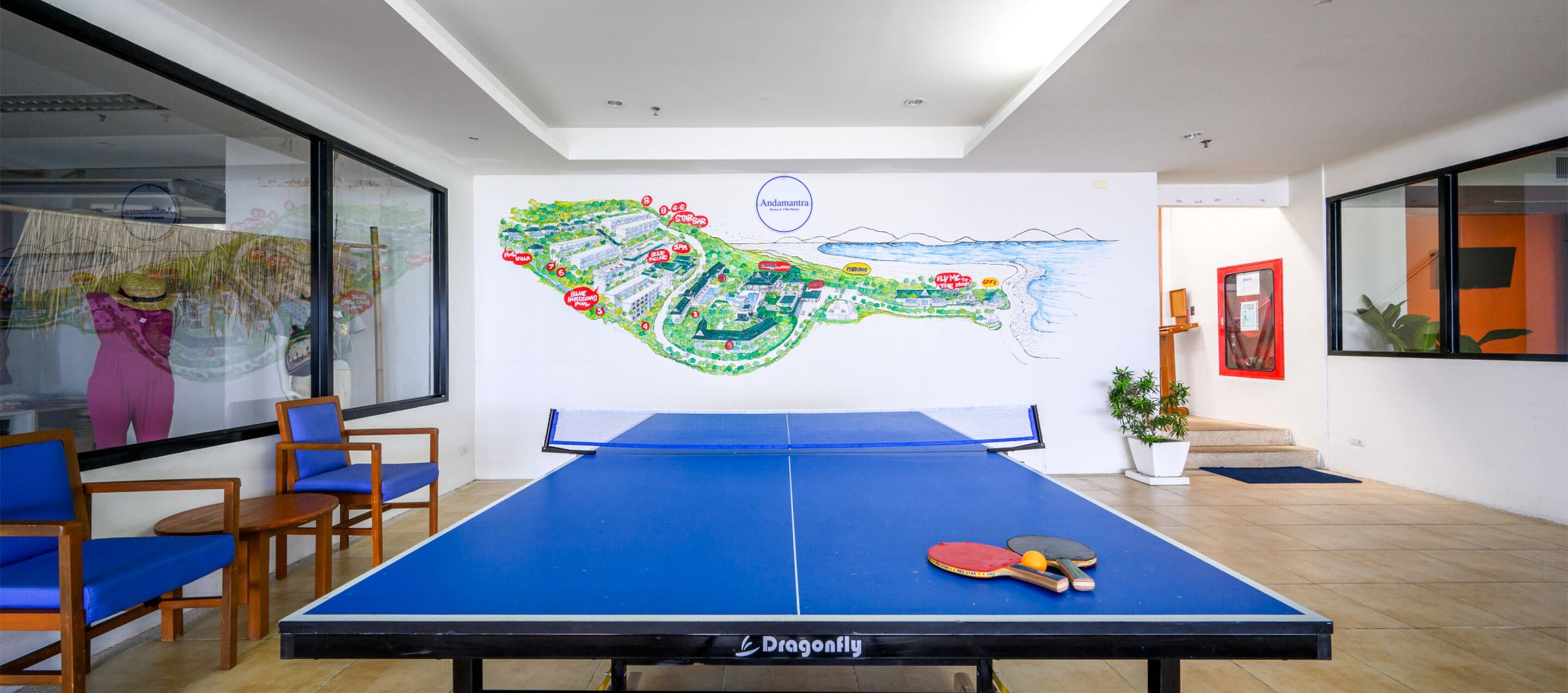 A ping pong table in a room with a large wall map, blue chairs, and a potted plant.