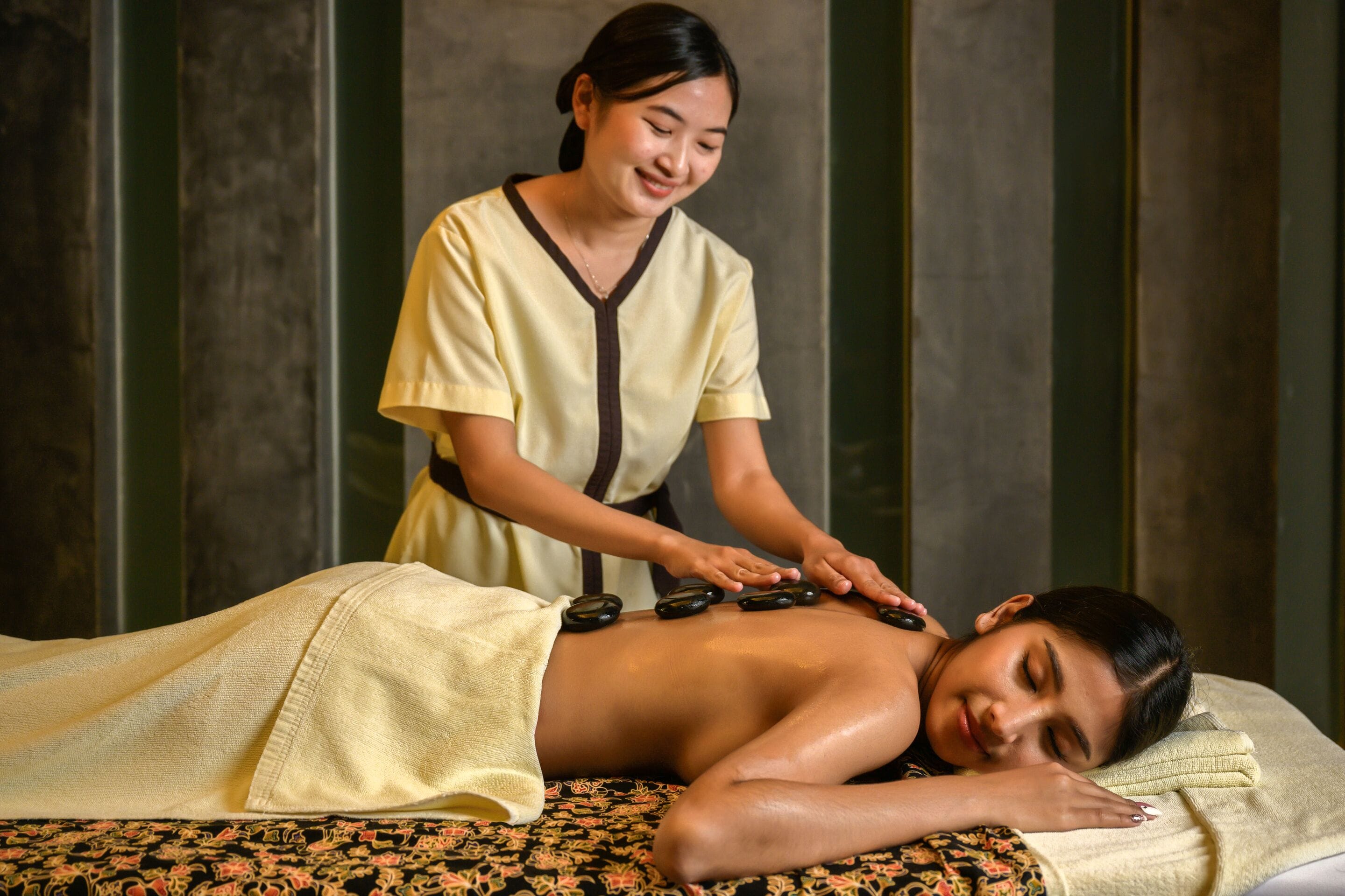 A massage therapy session in a spa setting with a client lying on a massage table, a therapist applying pressure with stones, and a patterned bedspread.