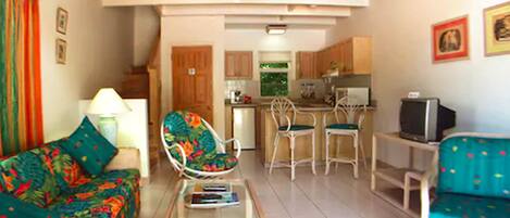 A living room with a sofa, a chair, a coffee table, and a television. There is a kitchen area with cabinets and a refrigerator.