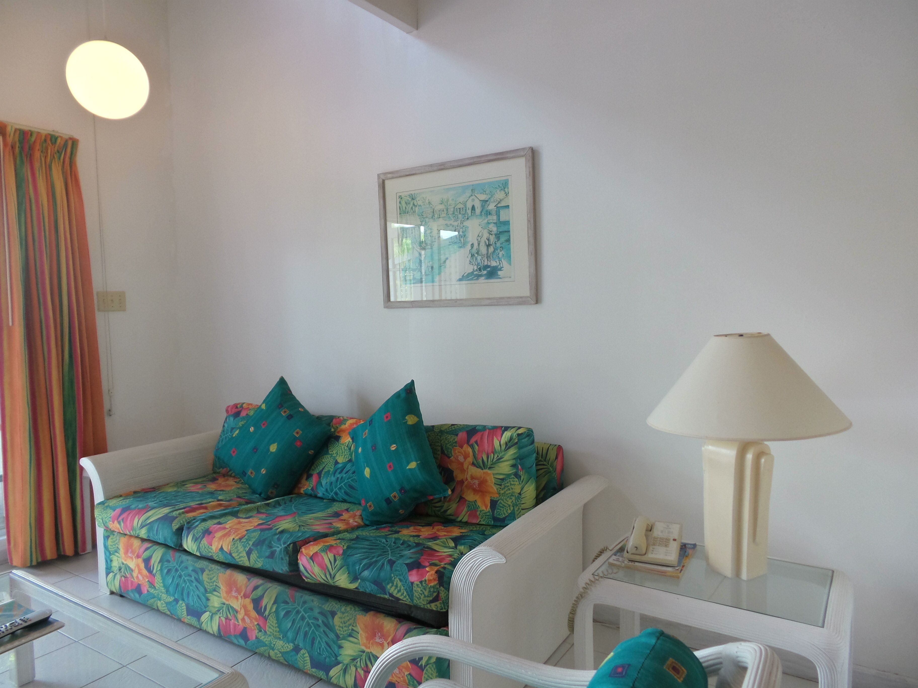 Un salon avec un canapé à motifs floraux, un fauteuil blanc, une table basse en verre, une lampe et un tableau encadré accroché au mur.