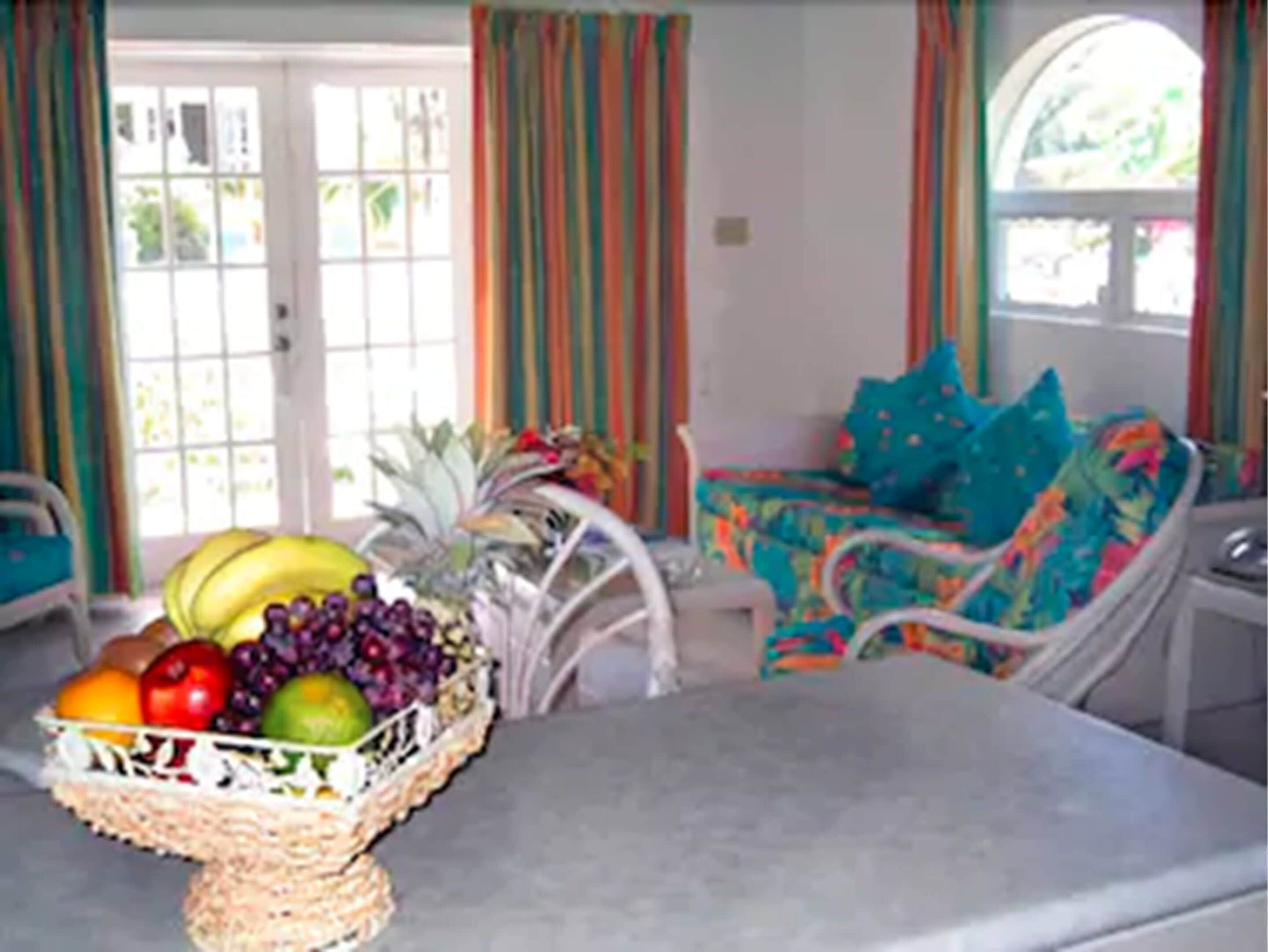 A room with a dining table, a wicker fruit basket, and a chair with a colorful cushion.