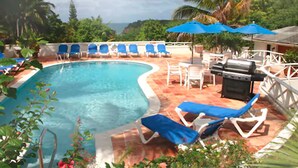 A pool area with lounge chairs and umbrellas, surrounded by greenery and a view of the ocean. - Dickenson Bay Cottages (St. John's)