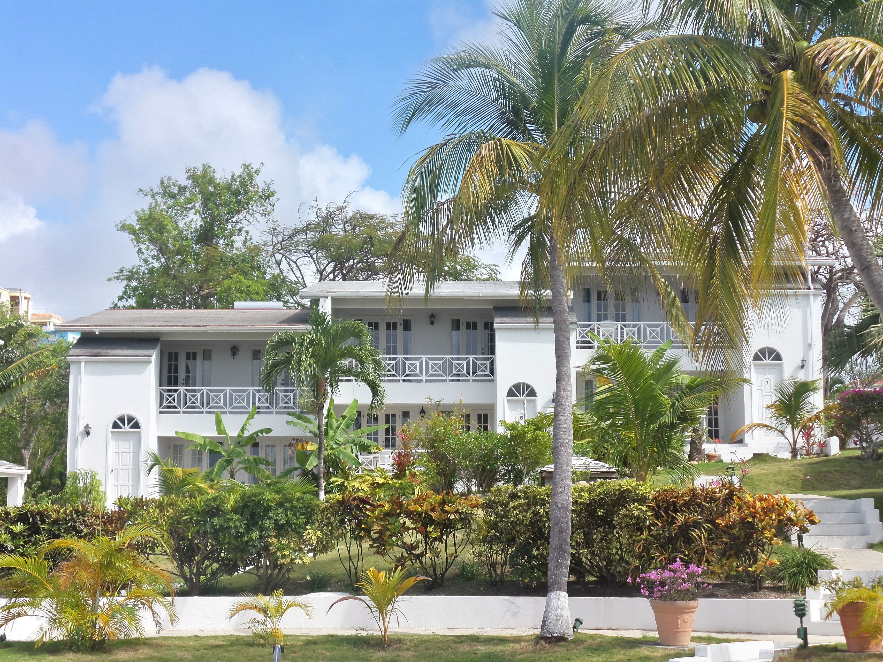 A white building with a balcony, surrounded by tropical plants and a clear sky.