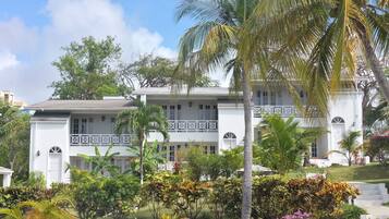 A white building with a balcony, surrounded by tropical plants and a clear sky.