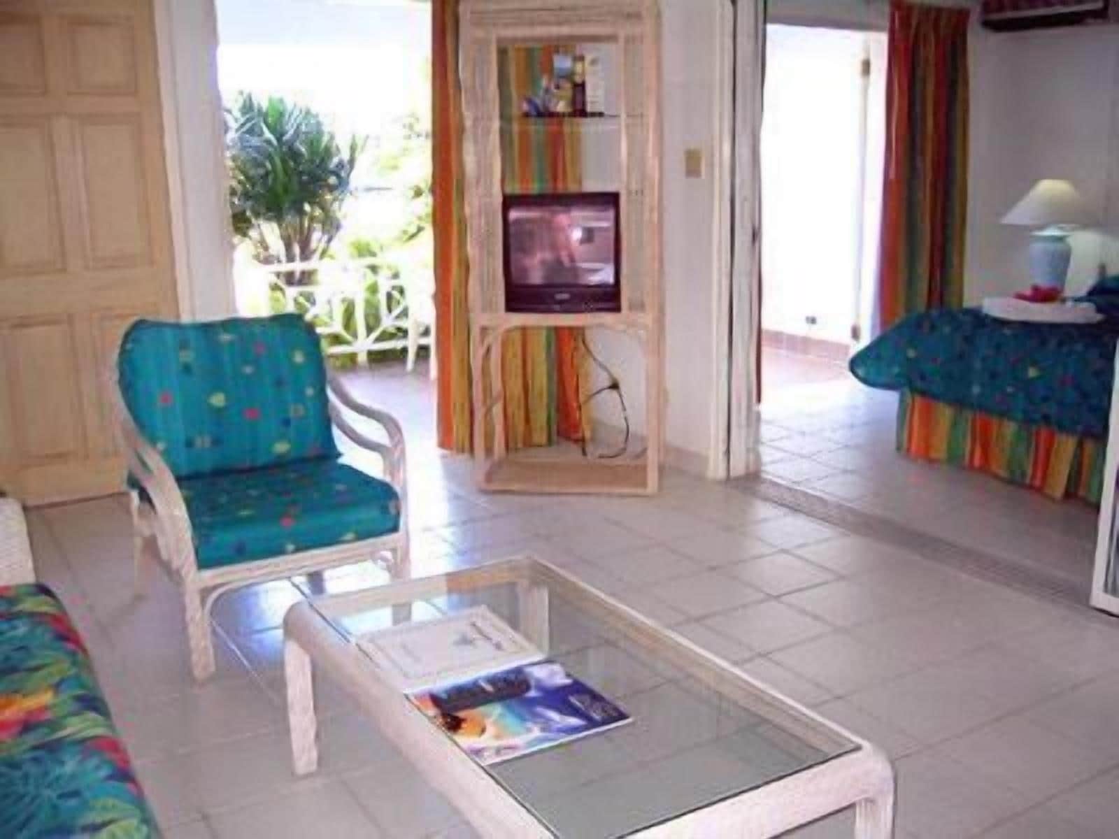A living room with a teal armchair, a glass coffee table with a magazine, and a television on a wooden stand.