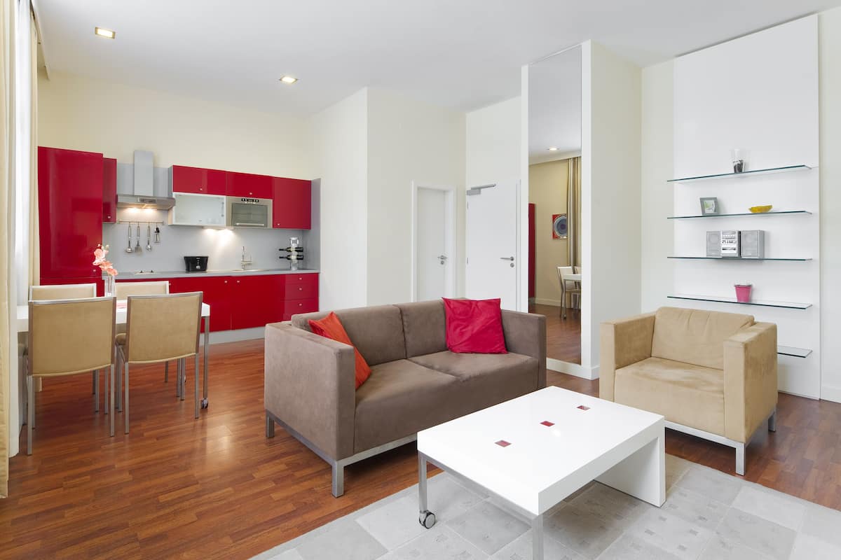 Una sala de estar moderna con un sofá, sillas y una mesa de centro. Se ve una cocina al fondo con armarios rojos y un microondas.