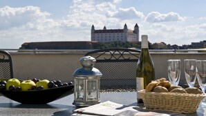Una mesa con un bol de fruta, un farol, una botella de vino y copas, y un castillo al fondo.