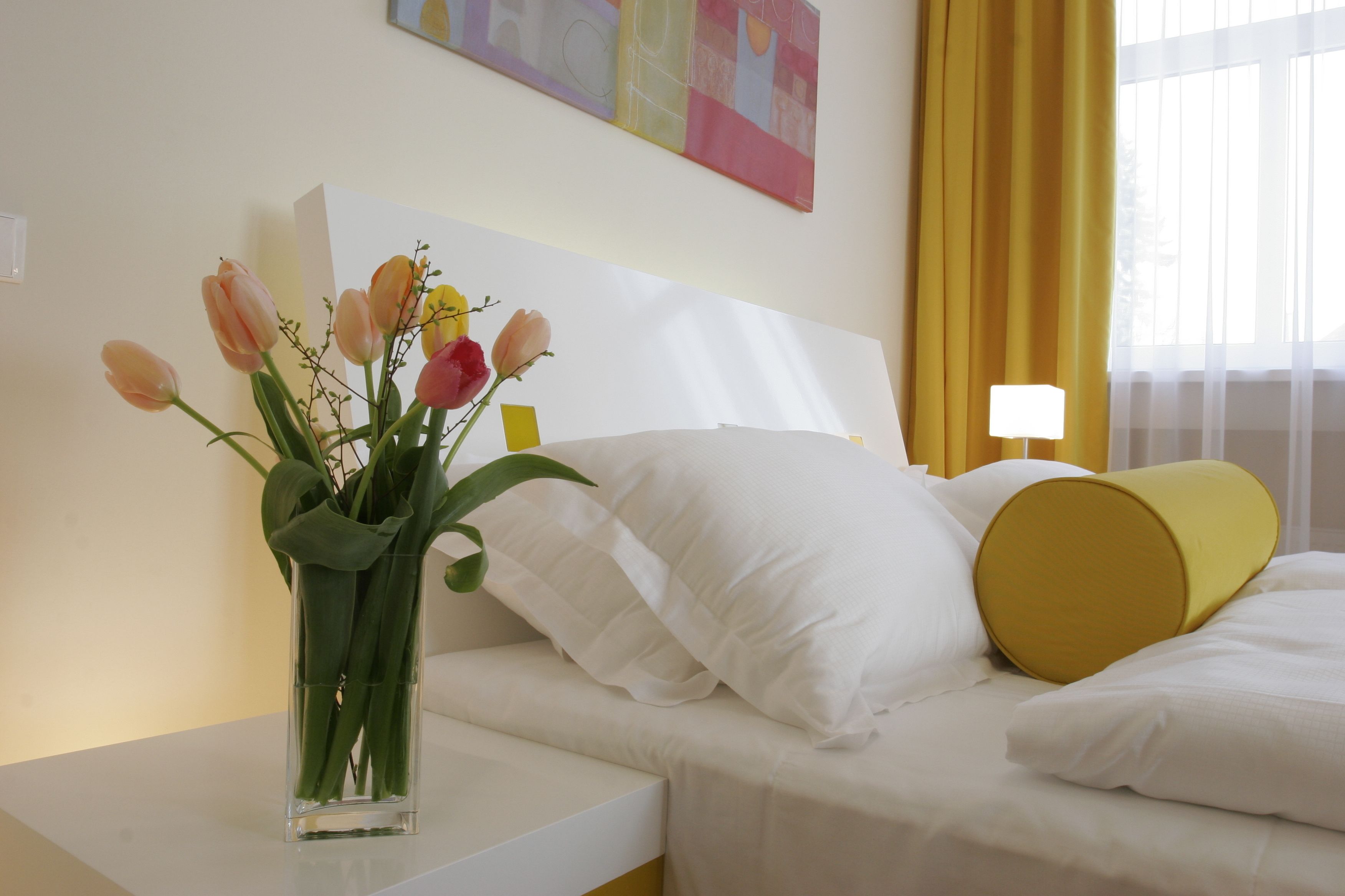 A hotel room with a bed, white pillows, a yellow accent pillow, a vase with flowers, and a painting on the wall.