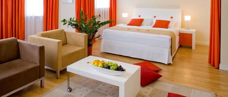 Una habitación de hotel moderna con una cama, un sofá, una mesita de centro con fruta y cortinas anaranjadas.