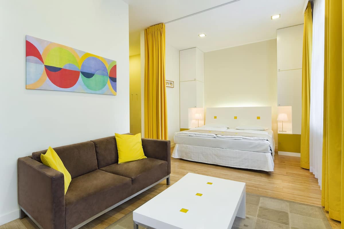 Una habitación de hotel moderna con un sofá marrón, una mesa de centro blanca, una cama con ropa de cama blanca y un cuadro abstracto colorido en la pared.