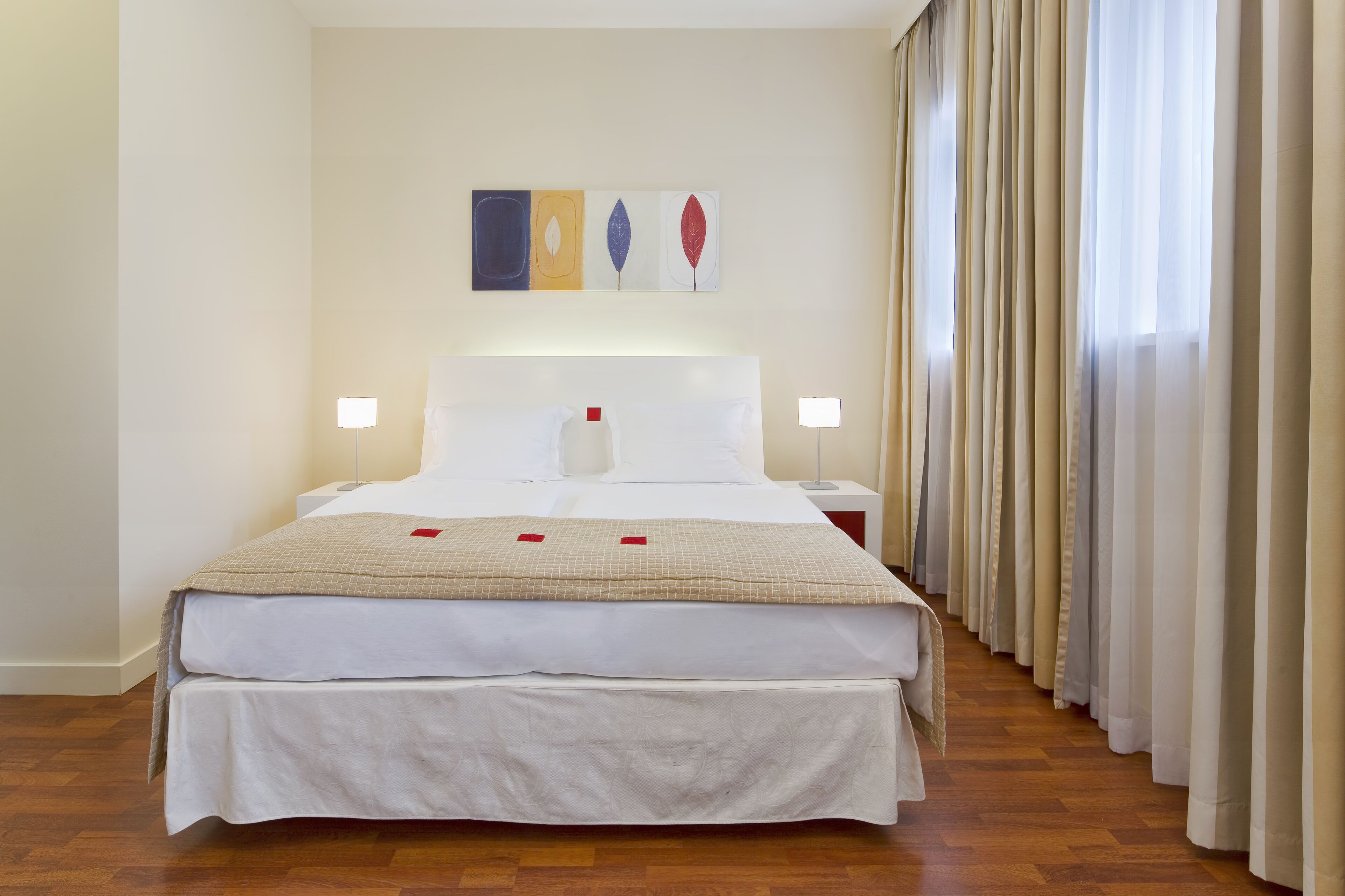 A hotel room with a large bed, two bedside tables, a wall-mounted artwork, and a window with curtains.