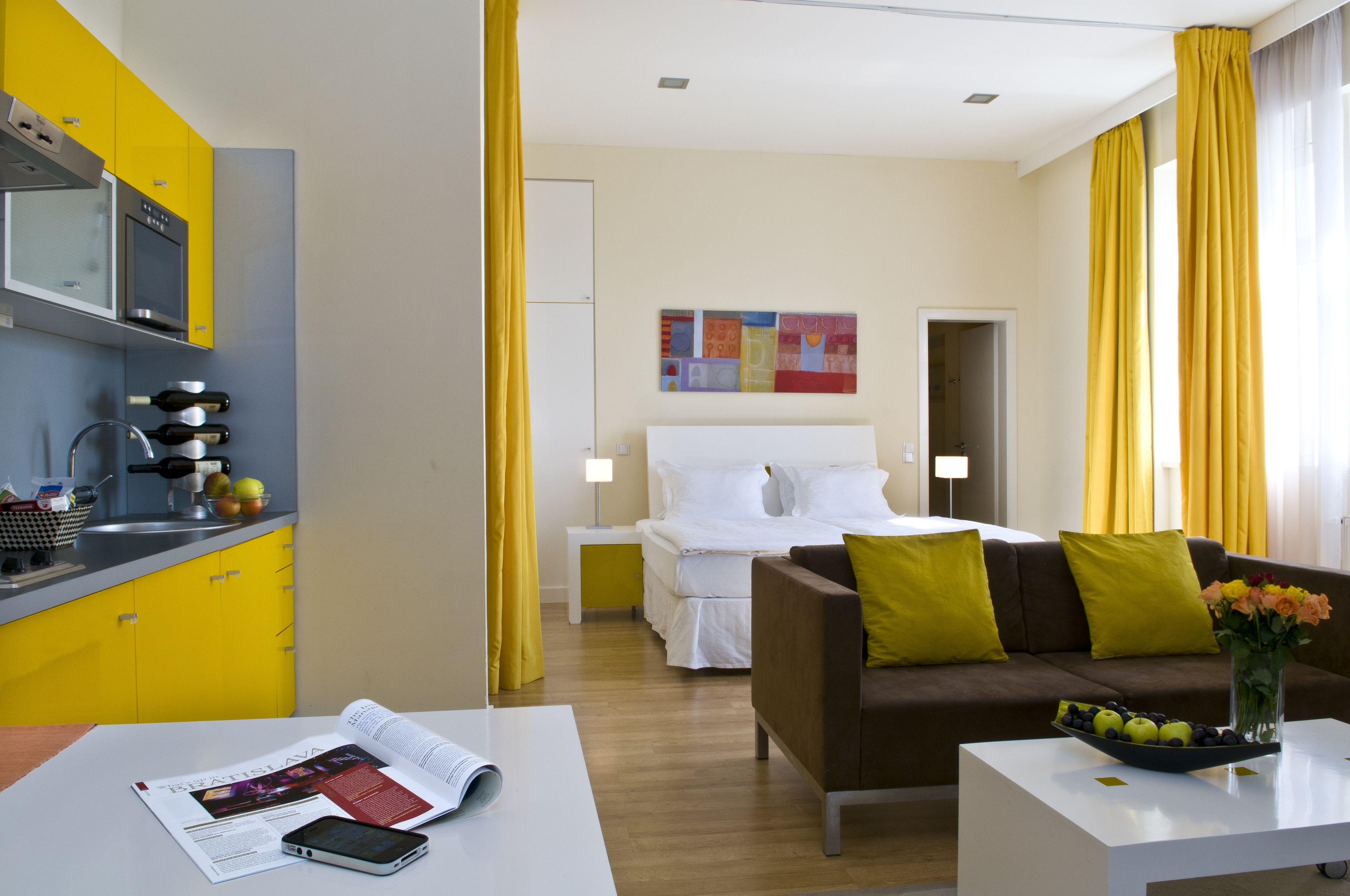 Ein modernes Hotelzimmer mit Küchenzeile, einem Bett mit weißer Bettwäsche, einem braunen Sofa mit gelben Kissen, einem weißen Couchtisch und einer Vase mit Blumen.