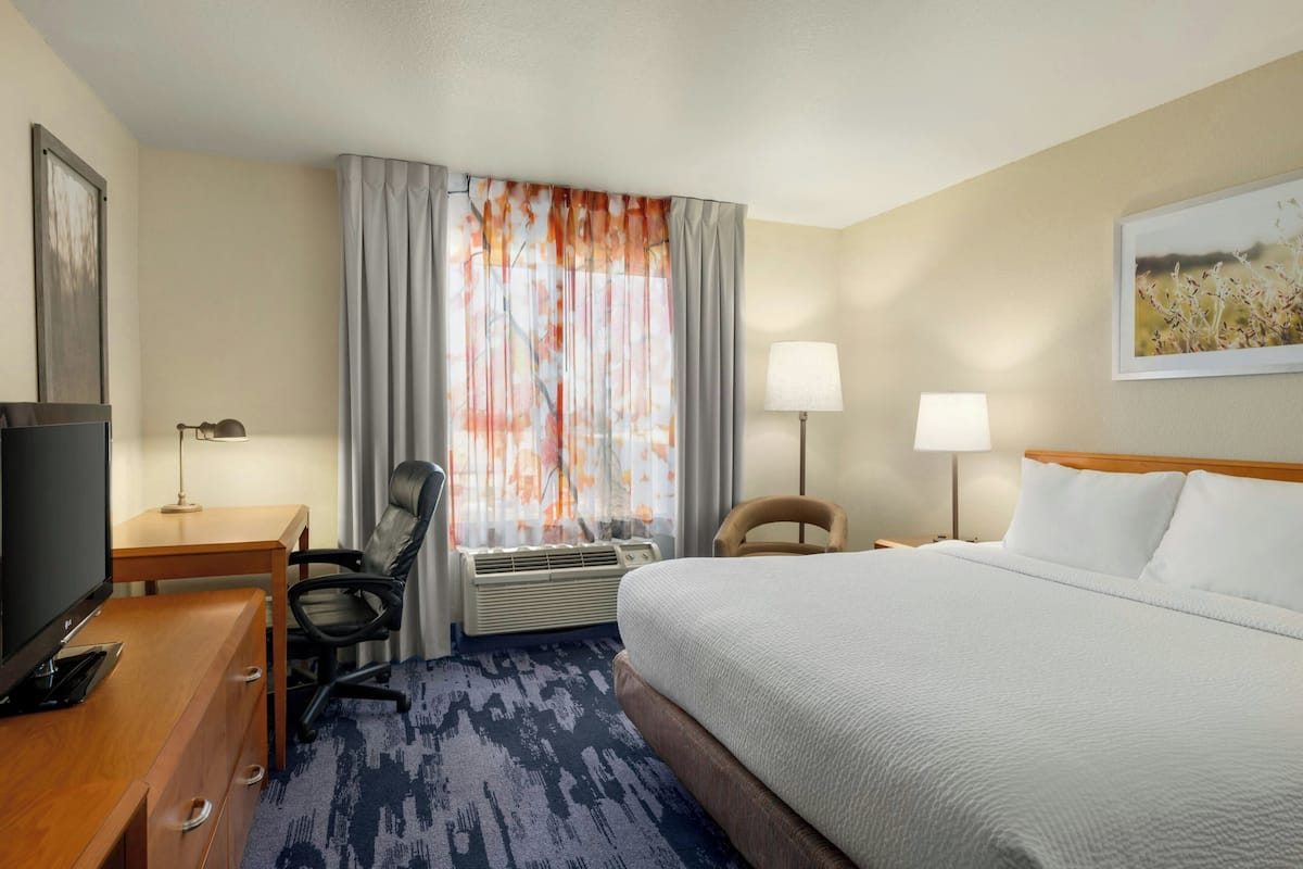 A hotel room with a bed, desk, chair, TV, air conditioning unit, and two lamps.