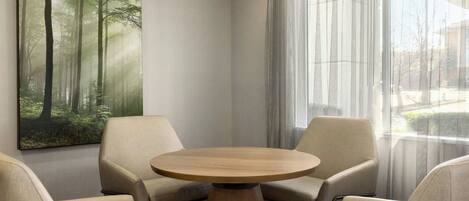 A corner room with a round wooden table and four beige armchairs. A large forest-themed painting is on the wall, and there is a window with sheer curtains.