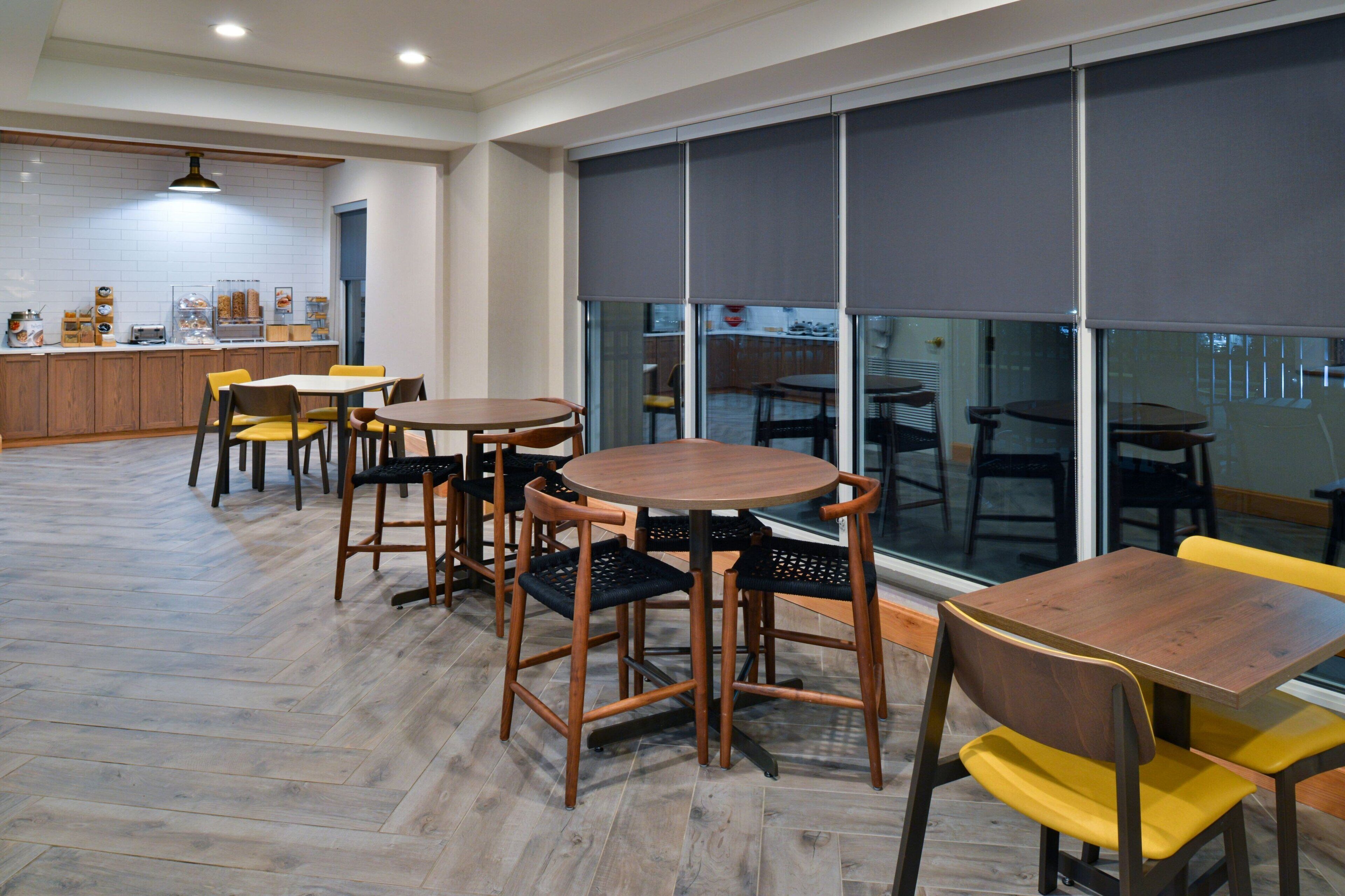A modern dining area with wooden tables and chairs, a kitchen in the background, and large windows with blinds.