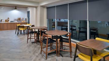 A modern dining area with wooden tables and chairs, a kitchen in the background, and large windows with blinds.