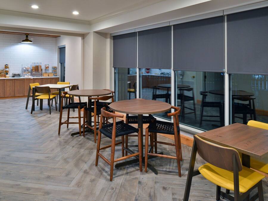 A modern dining area with wooden tables and chairs, a kitchen in the background, and large windows with blinds.