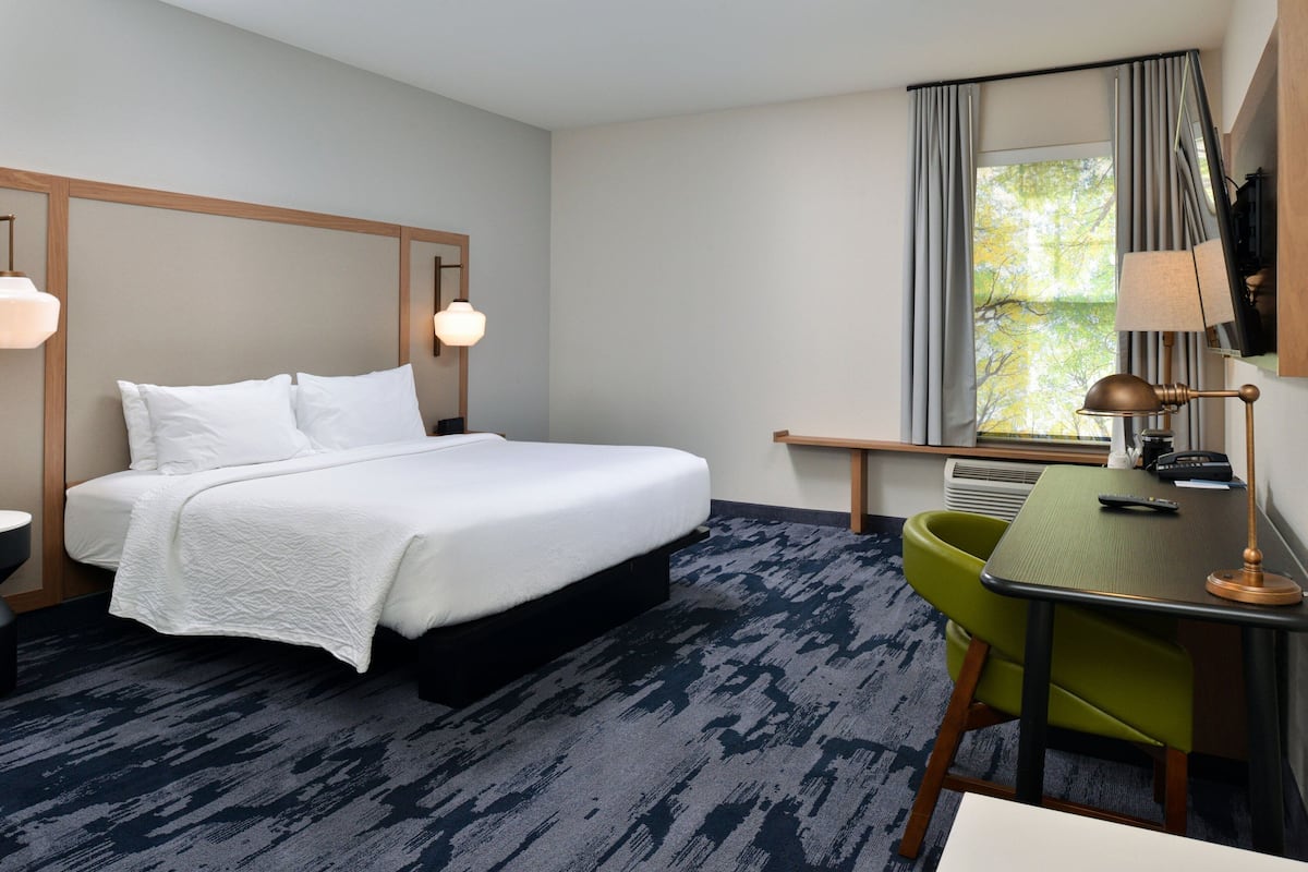 Une chambre d’hôtel avec un lit, un bureau, une chaise, une lampe et une fenêtre avec des rideaux.