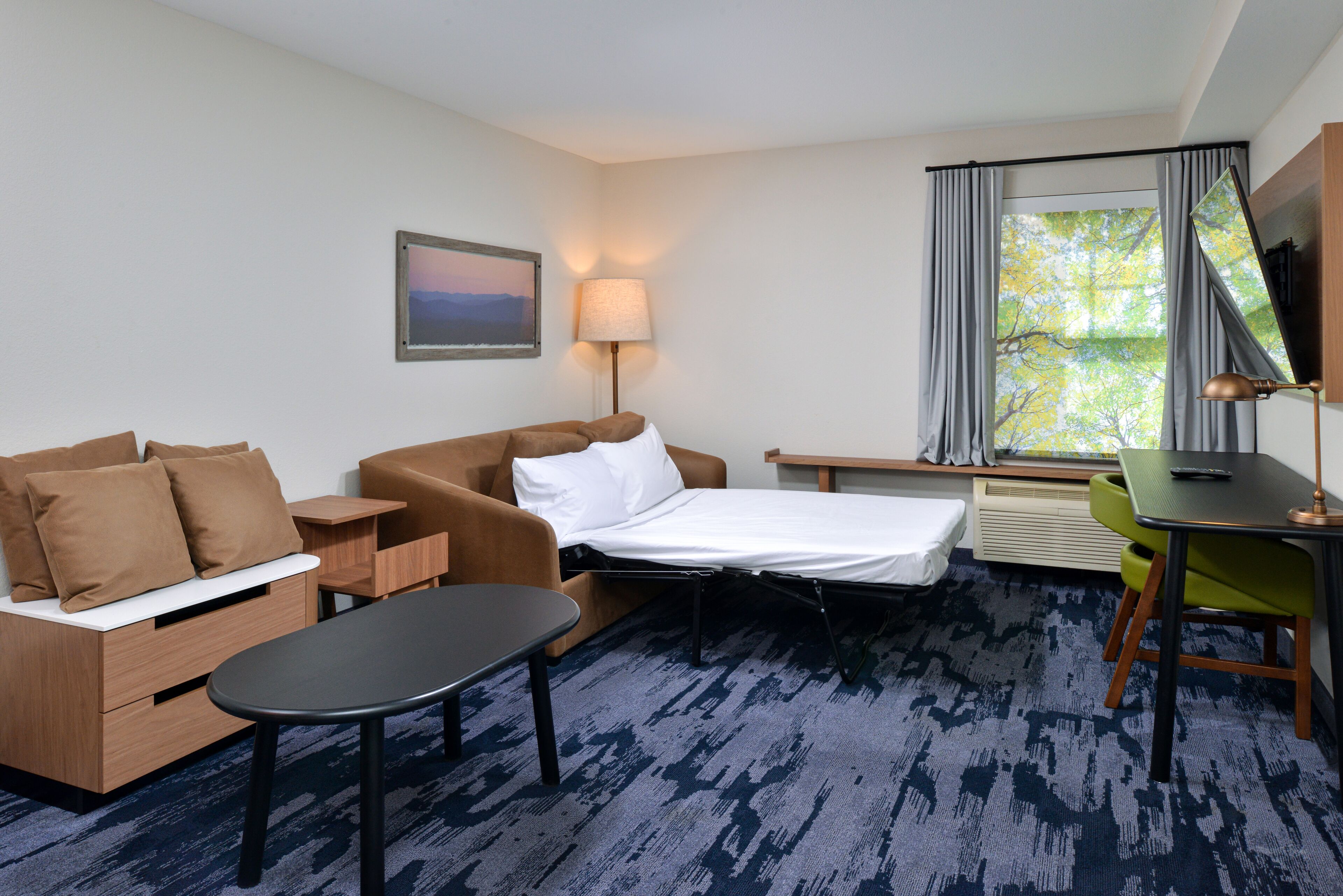 A hotel room with a bed, a sofa, a small table, a desk, a chair, a lamp, a window with curtains, and a painting on the wall.