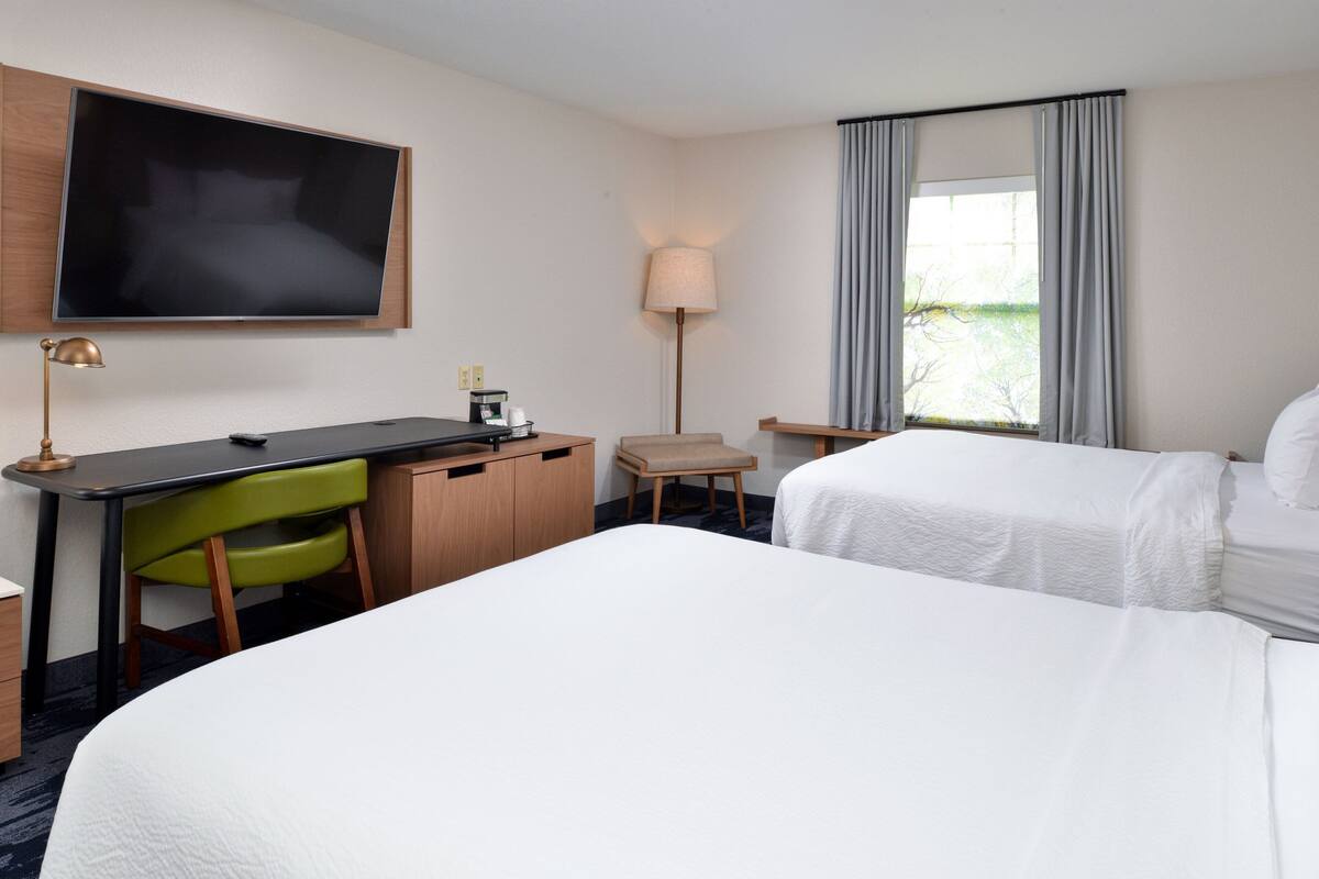 Une chambre d’hôtel avec un grand lit, un bureau avec une chaise, une télévision, une lampe et une fenêtre avec des rideaux.