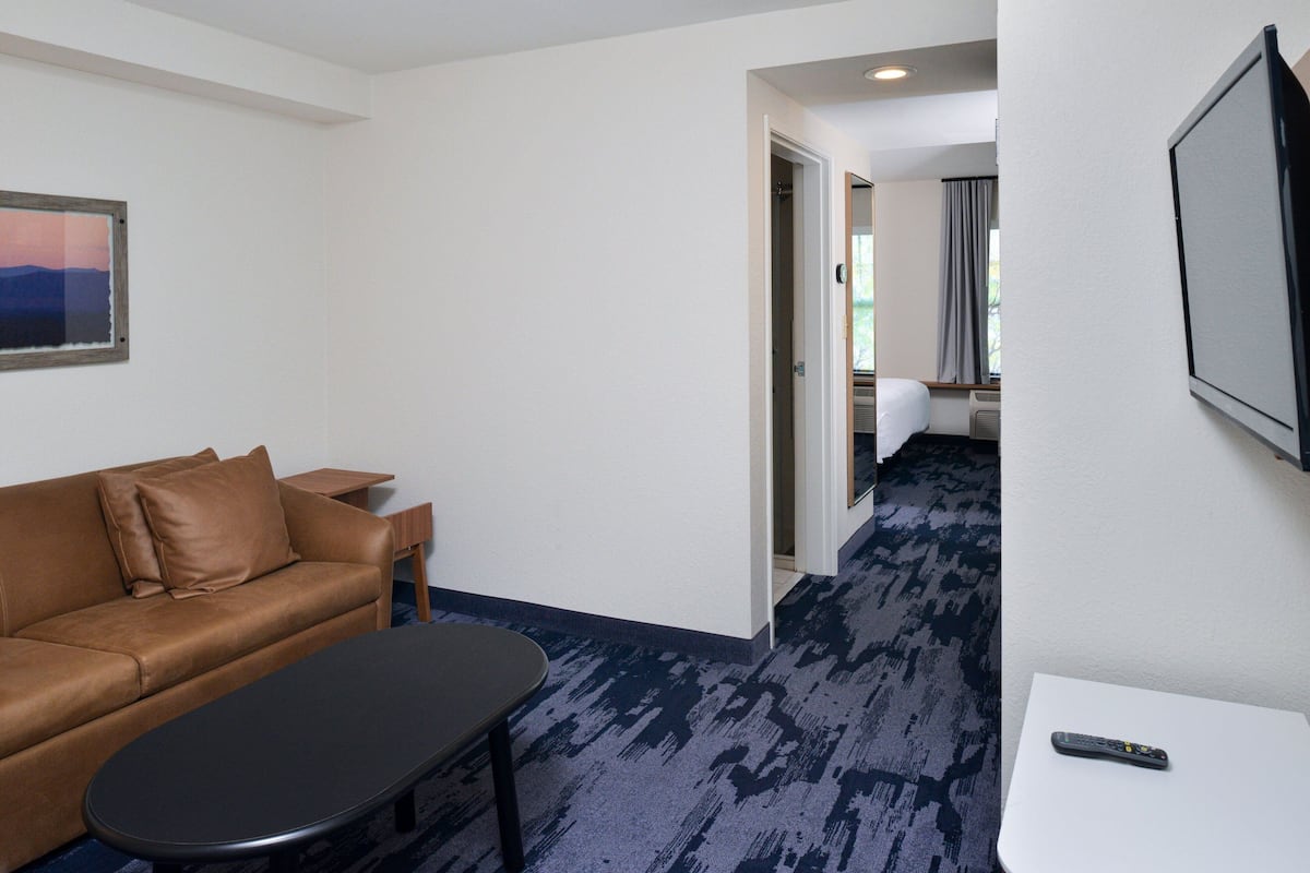 Une chambre d’hôtel avec un canapé en cuir marron, une table basse noire, un meuble TV blanc et une télécommande posée sur le meuble.