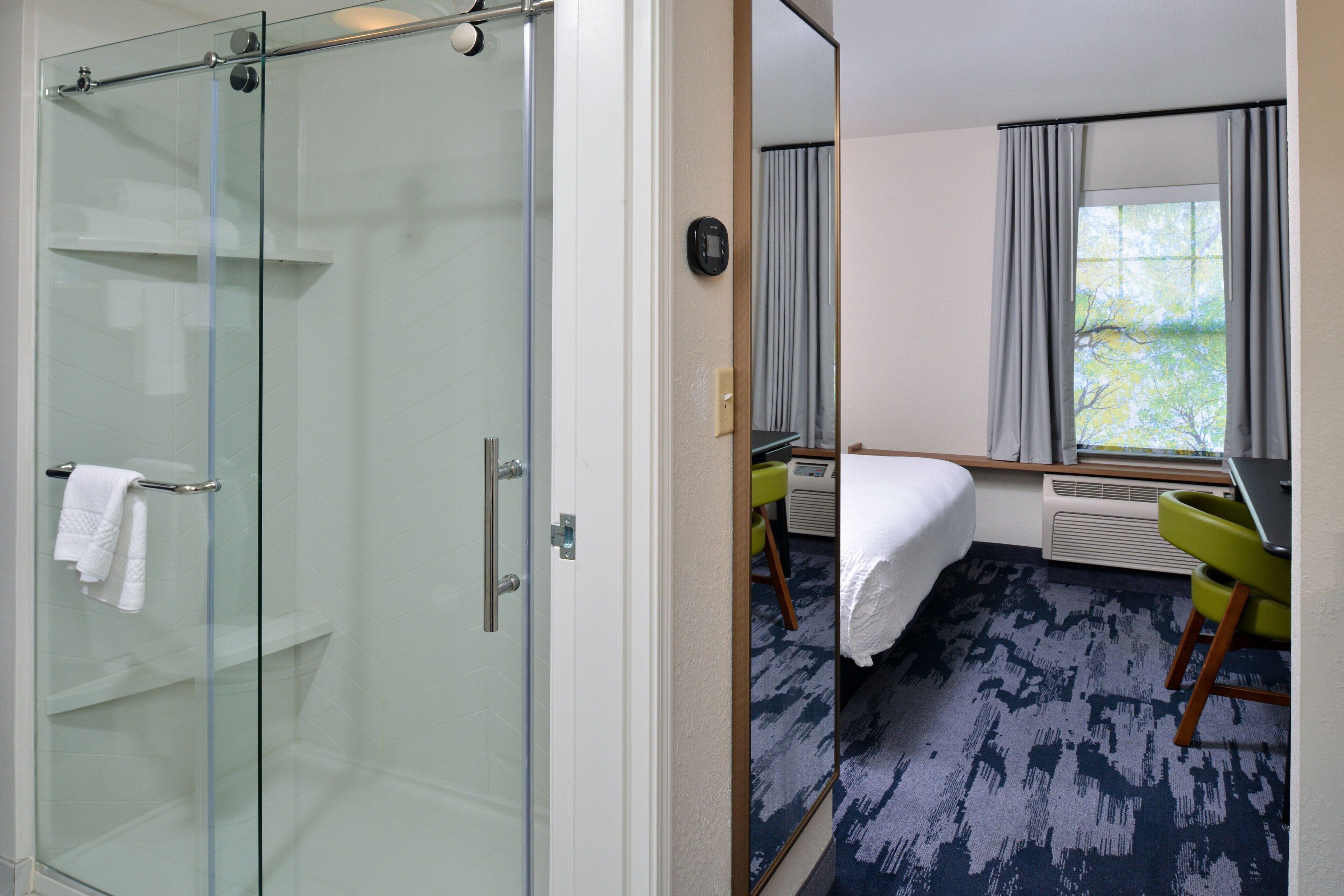 A hotel room with a glass-enclosed shower, a bed with a white comforter, a green chair, and a window with curtains.