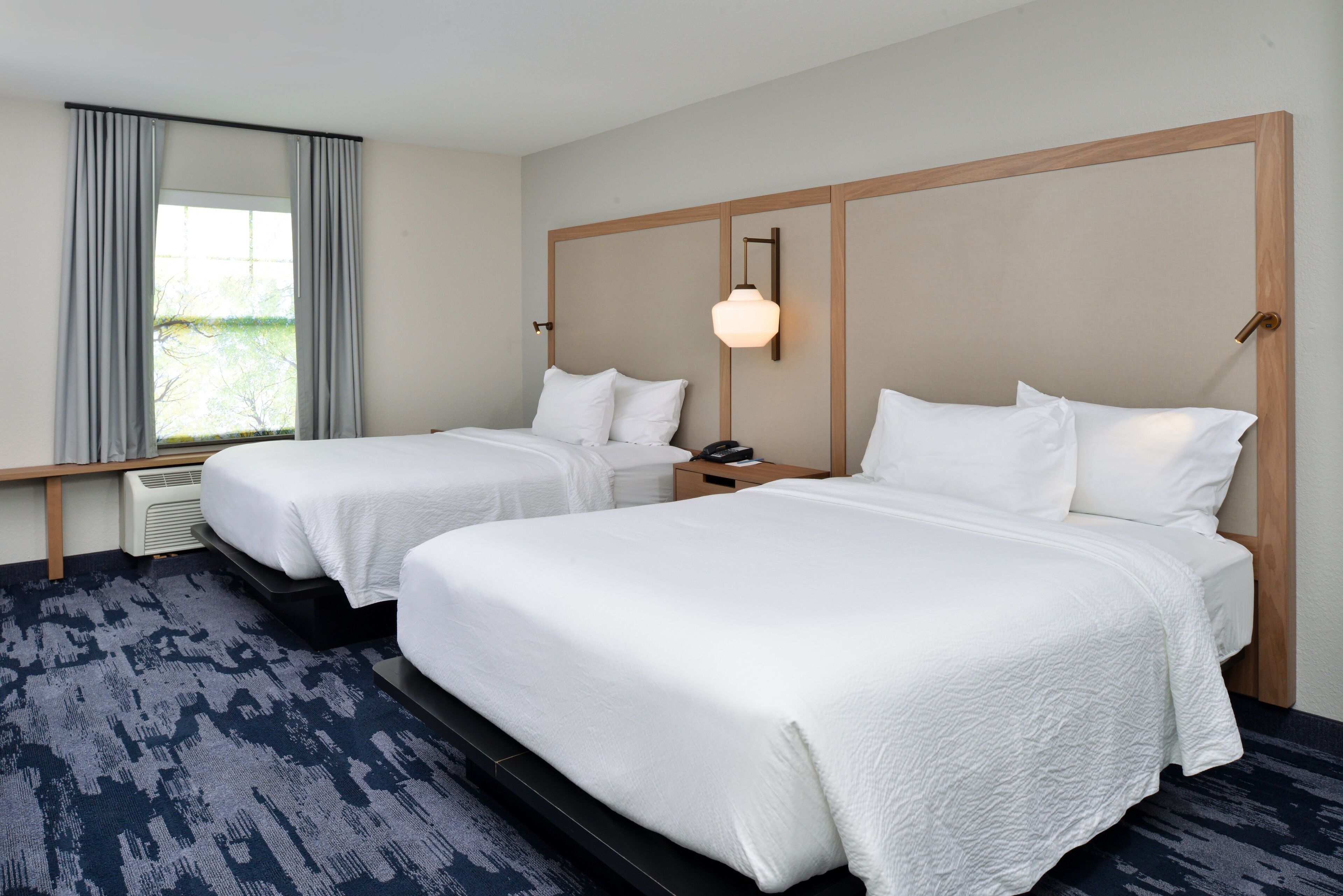 Une chambre d’hôtel avec deux lits, une grande fenêtre avec des rideaux, une lampe fixée au mur et un radiateur.