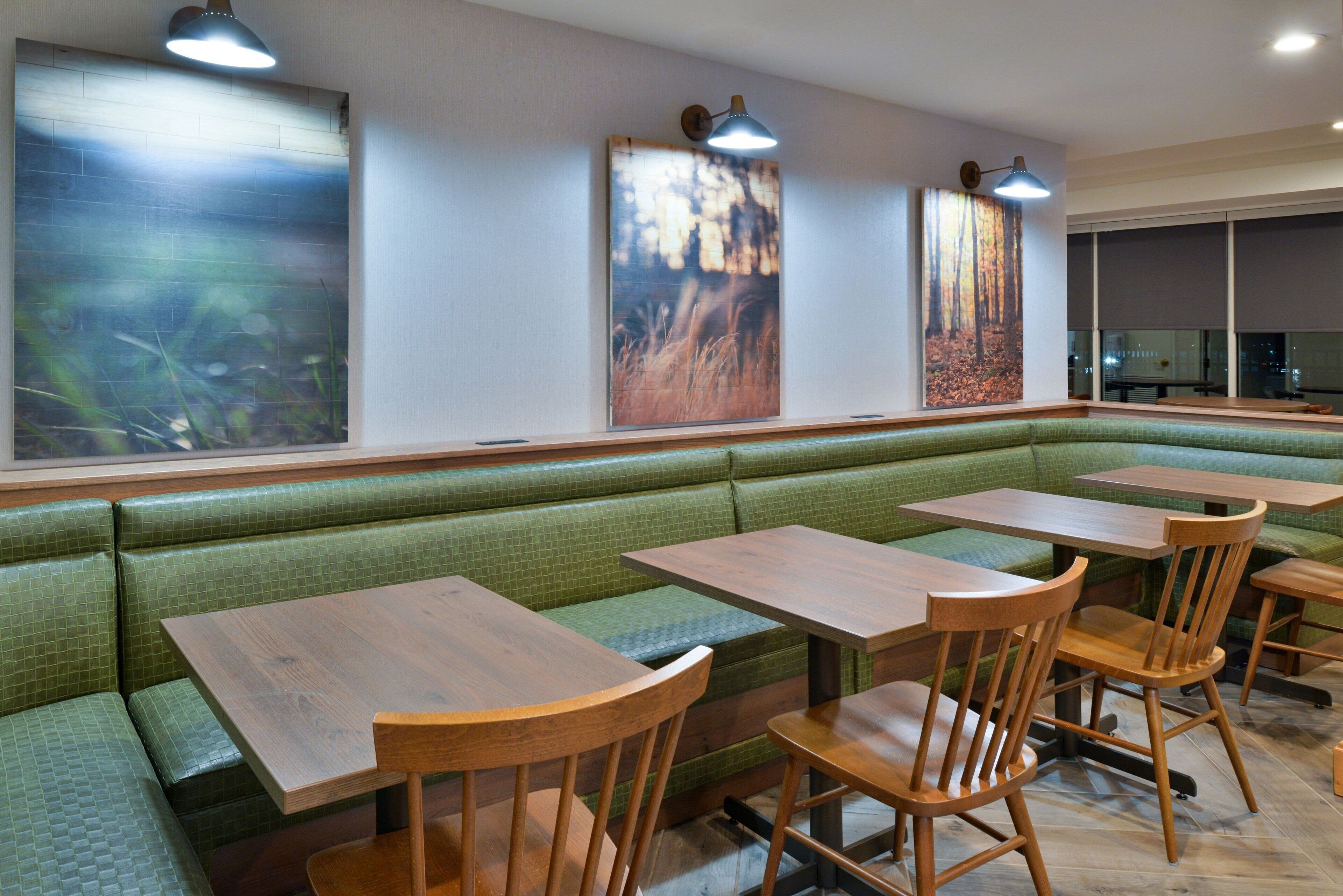 A restaurant with green cushioned booths, wooden tables, and chairs. There are wall-mounted light fixtures and large paintings on the walls.
