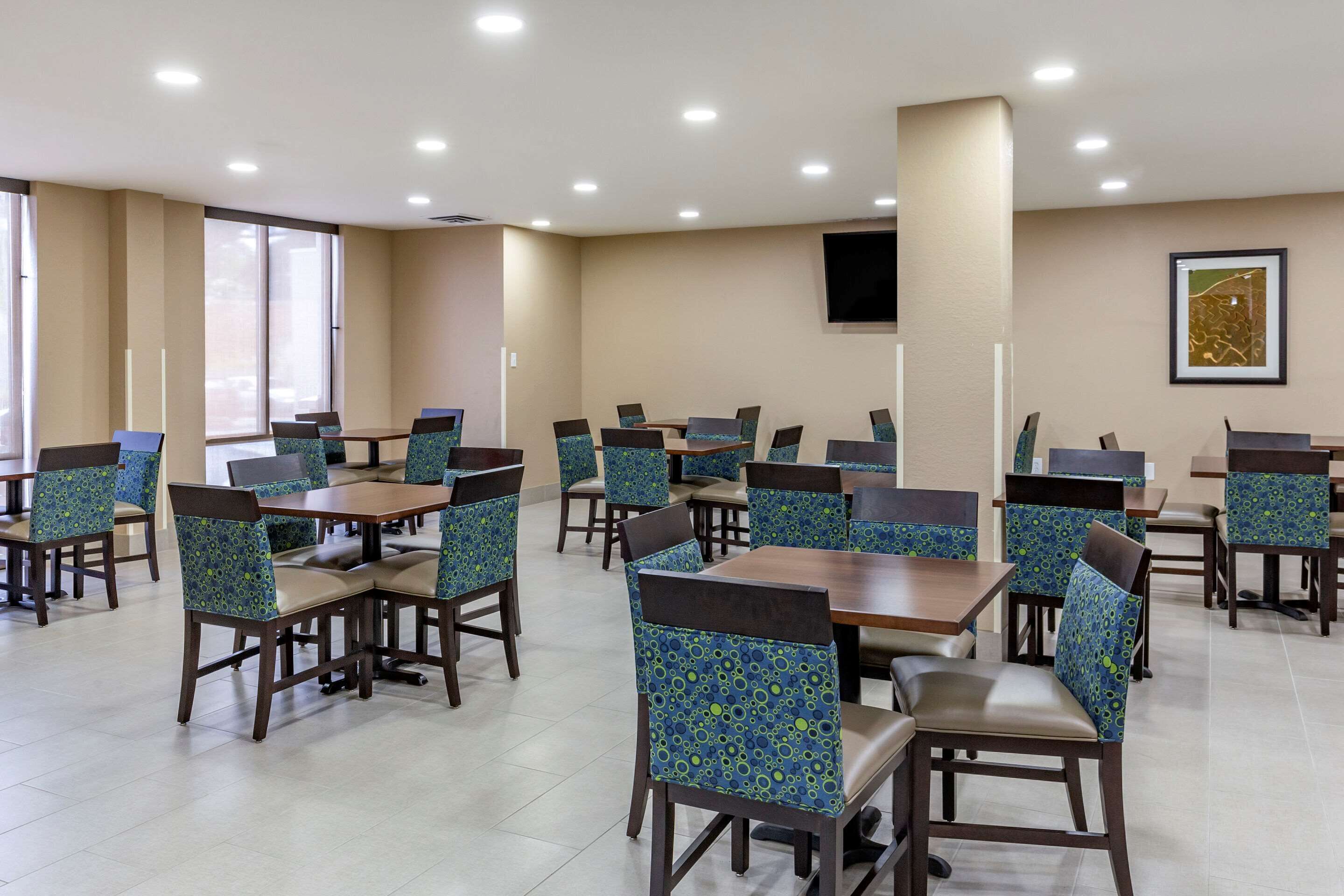 A hotel dining area with multiple tables and chairs, large windows, and a flat-screen TV on the wall.