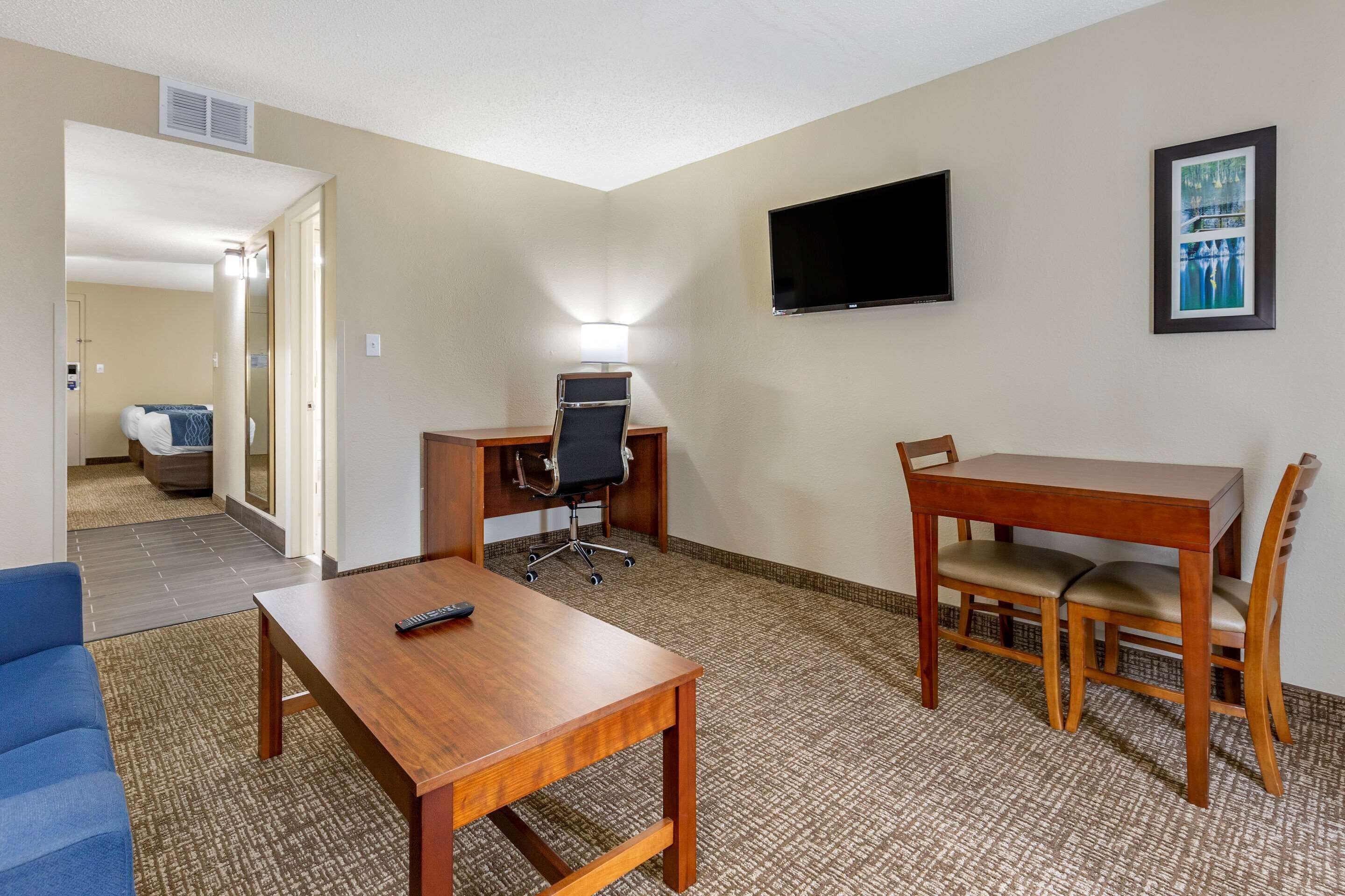 A hotel room with a wooden desk, a blue sofa, a dining table with chairs, a TV mounted on the wall, and a framed picture.