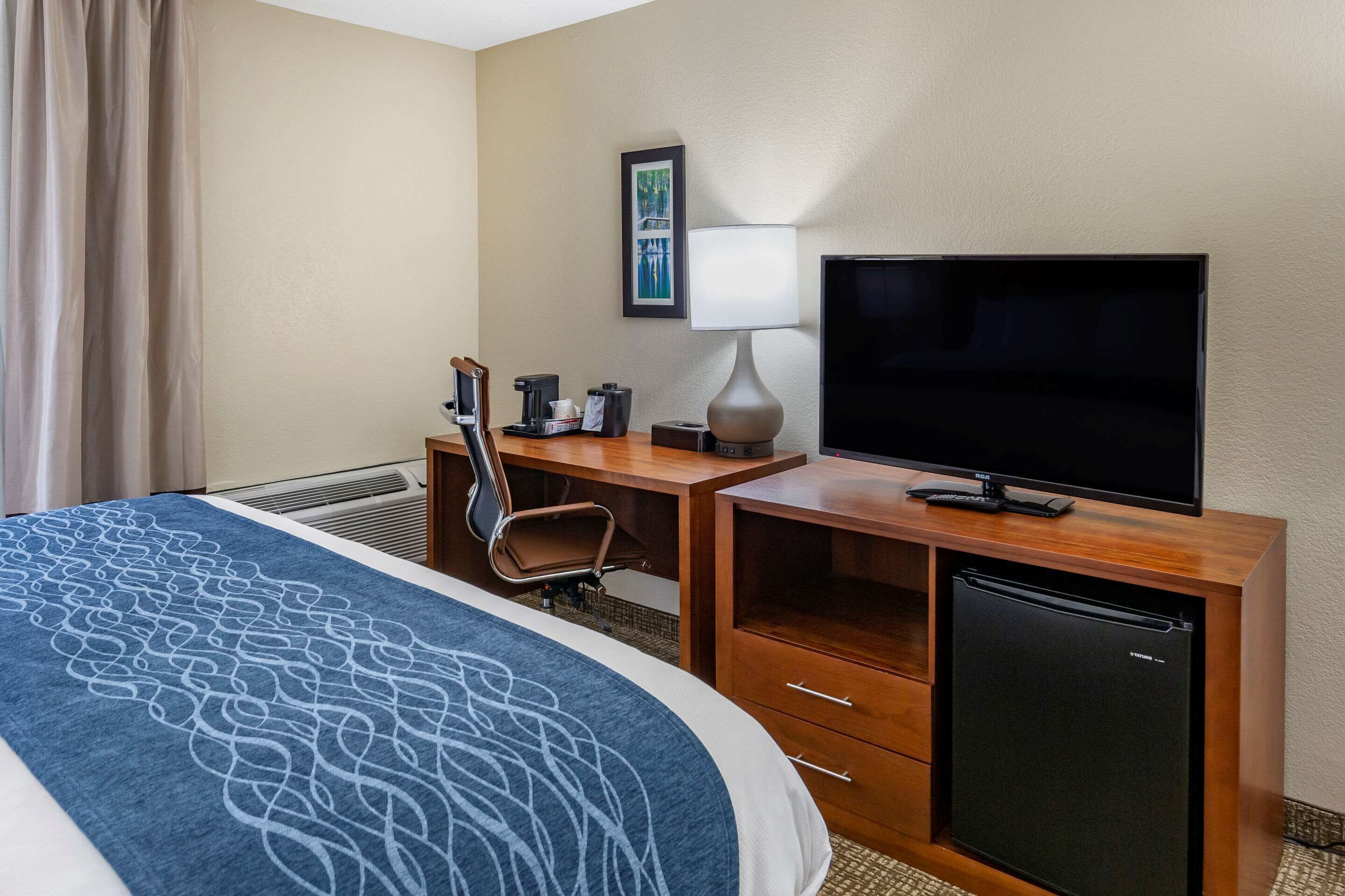 A hotel room with a bed, a desk with a television, a chair, a lamp, and a mini-fridge.