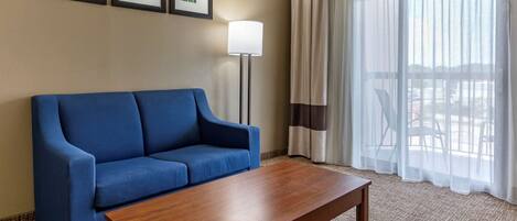 Habitación de hotel con sofá azul, mesa de centro de madera, lámpara de pie y ventana grande con cortinas translúcidas.