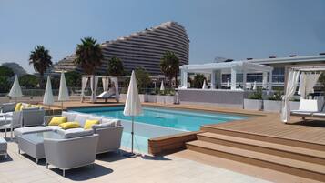 Un espace piscine aménagé sur le toit, avec des chaises longues, des parasols et une vue sur un bâtiment d’hôtel moderne.