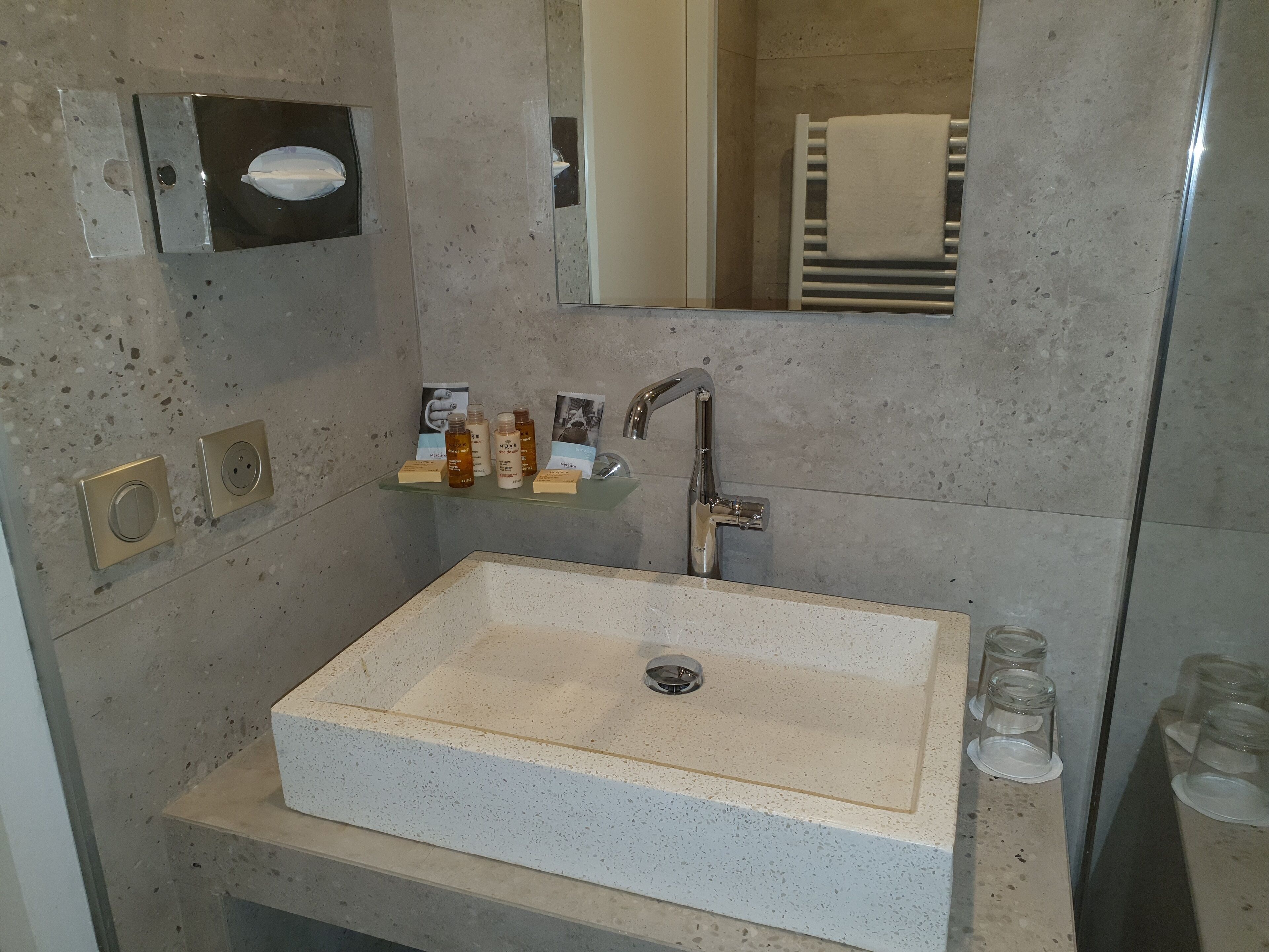Un baño con un lavamanos rectangular, un espejo, un perchero para toallas y un estante con artículos de aseo.